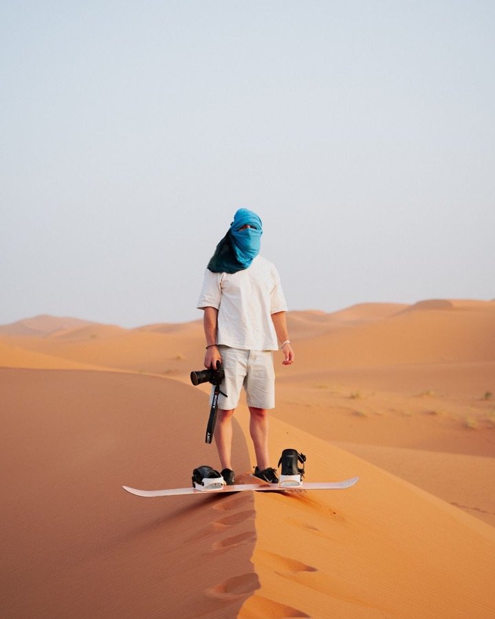 Jan Castany sobre una duna del desierto, con el rostro cubierto por un pañuelo azul, una cámara en la mano y una tabla de sandboard a los pies.